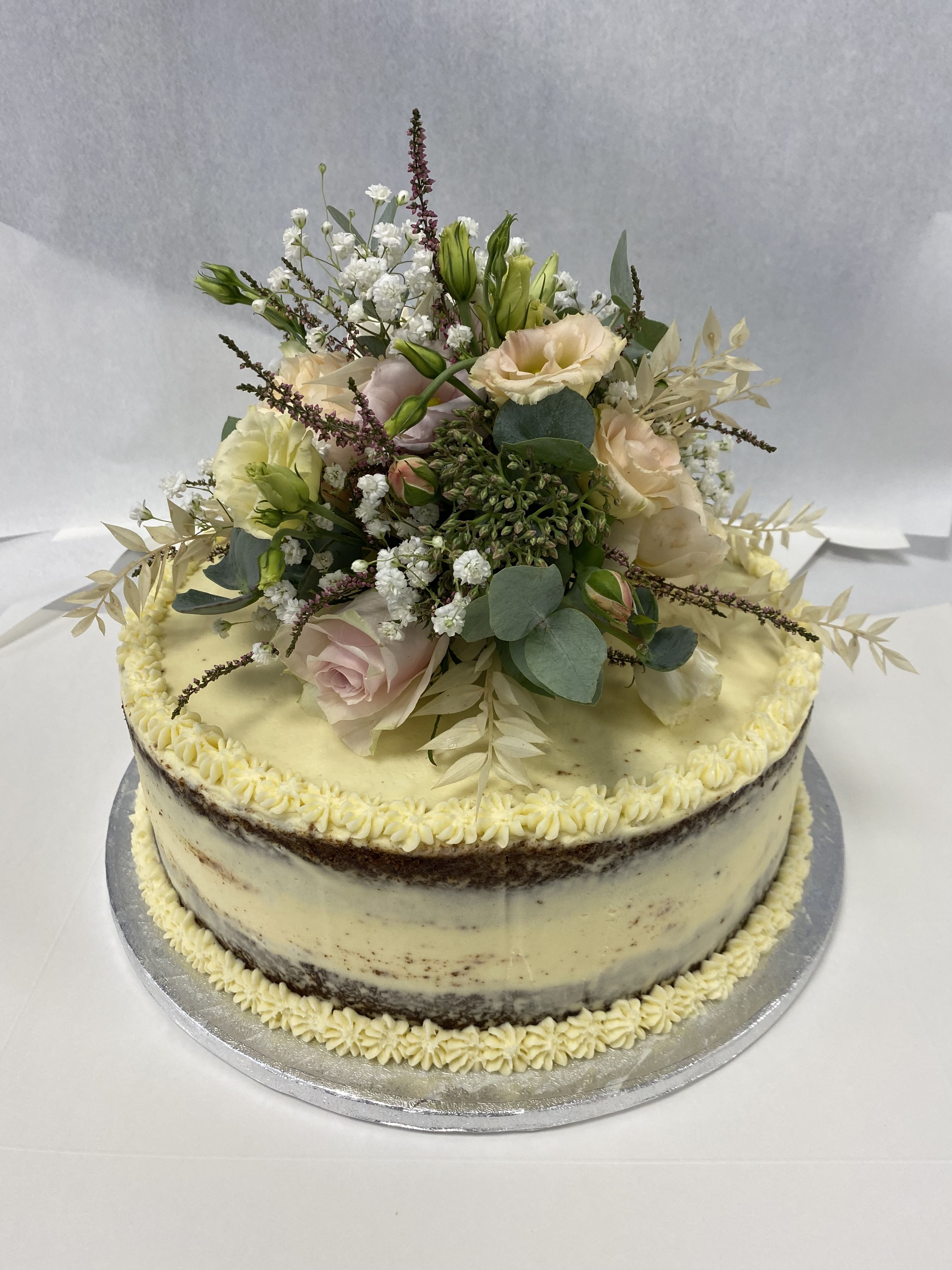 A two-tiered, semi-naked cake with light vanilla frosting is adorned with a bespoke arrangement of fresh flowers, including roses, baby's breath, and fern leaves. The stunning cake decoration sits on a silver cake board against a plain white background.